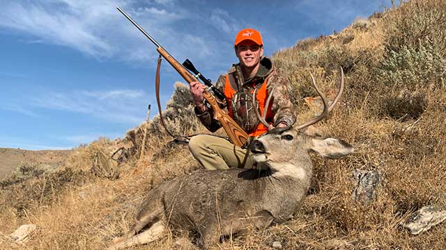 Young man, holding a rifle, crouched above a harvested deer