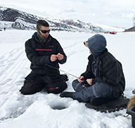 Chris Penne kneeling on the ice at Echo Reservoir, teaching a participant at an ice fishing clinic